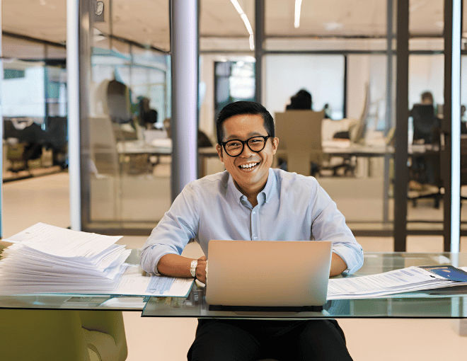 An Indonesian man, framed by bright office lighting, gazes happily at his laptop, with a backdrop of sleek office chairs and neatly arranged documents on a glass table.